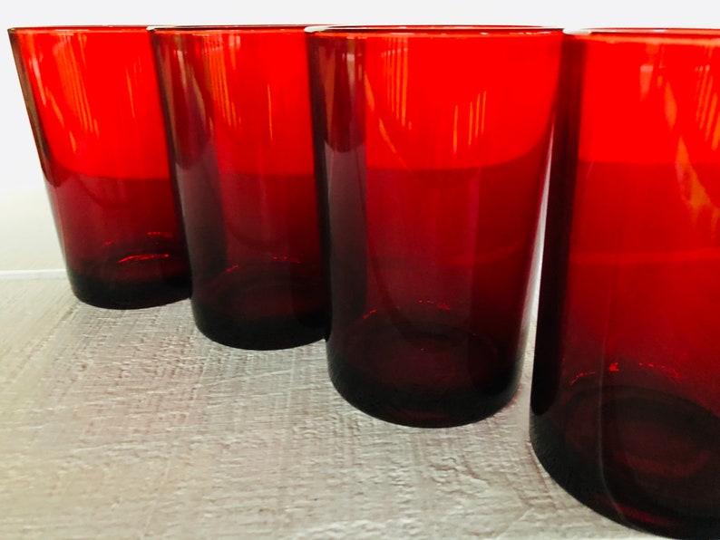 May include: Four tall, red-tinted drinking glasses. The glasses have a simple, cylindrical shape with a slightly tapered design. The color is a deep, rich red, and the glasses appear to be made of glass. The glasses are arranged in a row.