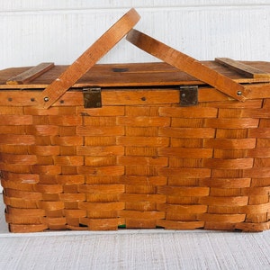 Puede incluir: Una cesta de picnic de madera vintage con tapa abatible y dise&ntilde;o tejido. La cesta tiene dos cierres met&aacute;licos y un asa de madera marr&oacute;n.