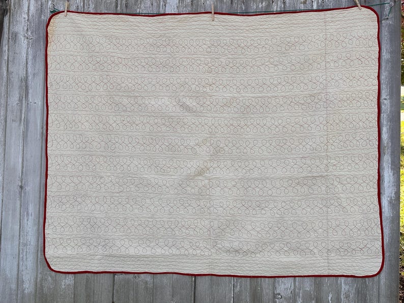 Peut inclure: Une courtepointe rectangulaire cr&egrave;me avec une bordure rouge, suspendue &agrave; une corde &agrave; linge. La courtepointe pr&eacute;sente un motif r&eacute;p&eacute;titif de boucles interconnect&eacute;es de couleur cr&egrave;me plus claire, cr&eacute;ant un effet textur&eacute;. Le design g&eacute;n&eacute;ral est simple et classique.