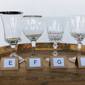 May include: Four clear glass wine glasses with silver rims on a wooden tray. The glasses are labeled E, F, G, and H.