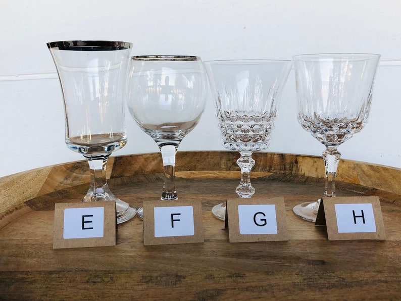 May include: Four clear glass wine glasses with silver rims on a wooden tray. The glasses are labeled E, F, G, and H.