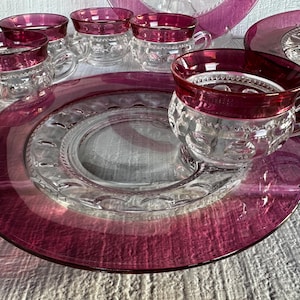 May include: A set of vintage clear glass teacups and saucers with a ruby red rim. The cups and saucers have a detailed, textured design. The set includes multiple cups and plates, perfect for serving tea or coffee.