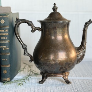 May include: A tarnished silver-plated teapot featuring an ornate curved handle, a decorative spout, and a lid with a finial top. The teapot stands on three small feet and has a bulbous body with visible signs of oxidation and dark patina.