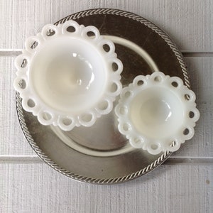 May include: Two white milk glass bowls with scalloped edges sit on a silver charger. The bowls are different sizes and have a vintage look.