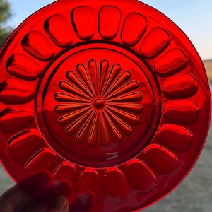 May include: A vibrant red glass plate with a detailed floral center design and a scalloped edge. The plate's glossy surface reflects light, highlighting its intricate patterns. A decorative piece for serving or display.
