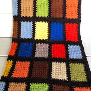 May include: A colorful crocheted blanket with squares in various colors, including red, orange, yellow, green, blue, brown, and beige. The squares are arranged in a grid pattern and are separated by black borders.