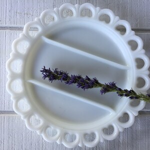 May include: White milk glass divided serving dish with a scalloped edge. The dish has three sections and is decorated with a delicate lace pattern. A sprig of purple flowers is resting in the dish.