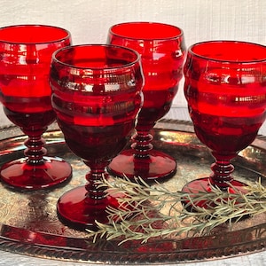 May include: Four ruby red wine glasses with a ribbed design are displayed on a silver serving tray. The glasses have a classic goblet shape with a round base and a decorative stem. The tray has an ornate, embossed pattern.
