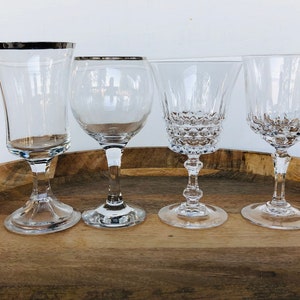 May include: Four clear glass wine glasses on a wooden surface. The glasses have different shapes and styles, including a tall, slender glass with a silver rim, a wide-mouthed glass with a short stem, and two glasses with a more traditional shape and a longer stem.