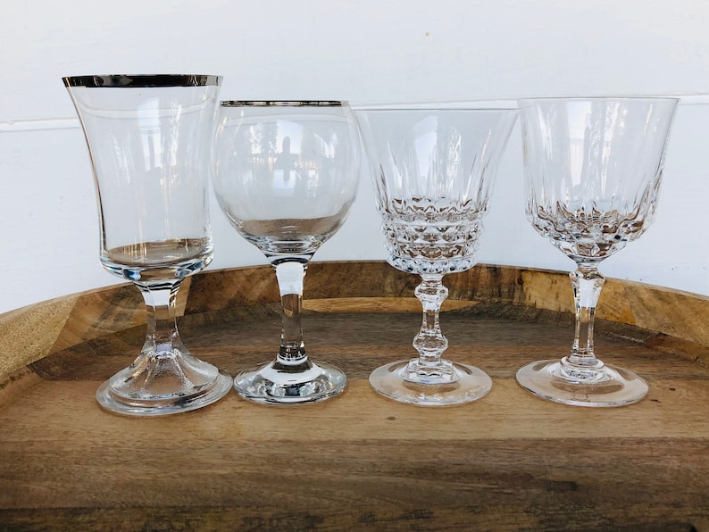 May include: Four clear glass wine glasses on a wooden surface. The glasses have different shapes and styles, including a tall, slender glass with a silver rim, a wide-mouthed glass with a short stem, and two glasses with a more traditional shape and a longer stem.