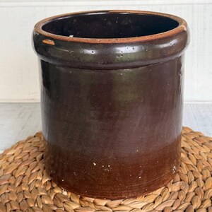 May include: A cylindrical stoneware crock featuring a deep, dark brown glazed finish. The vessel has a thick, rounded rim at the top with a smooth, glossy texture throughout the exterior surface, showing subtle variations in the brown color tone.