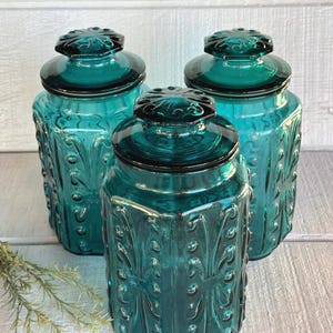 May include: Three teal glass jars with lids. The jars have a square shape with embossed floral designs. The lids are round with a decorative knob. The jars are arranged on a white wooden surface.