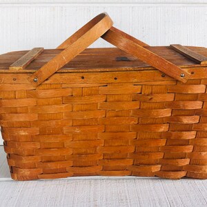 Puede incluir: Una cesta de picnic de madera vintage con un dise&ntilde;o tejido y una tapa con bisagras. La cesta tiene un asa de madera marr&oacute;n y est&aacute; hecha de madera natural.