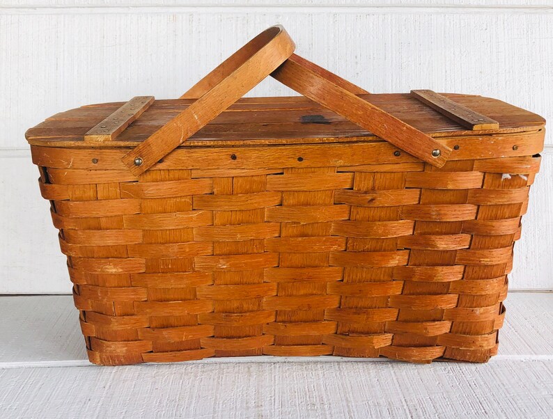 Puede incluir: Una cesta de picnic de madera vintage con un dise&ntilde;o tejido y una tapa con bisagras. La cesta tiene un asa de madera marr&oacute;n y est&aacute; hecha de madera natural.