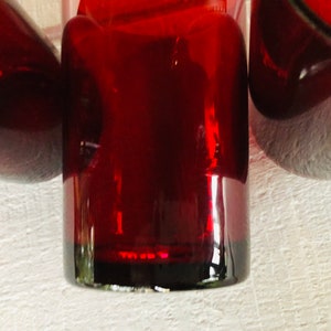 May include: Close-up of three ruby red glass objects. The objects have a glossy finish and are arranged closely together. The lighting creates highlights and shadows, emphasizing the shapes and the rich color of the glass.