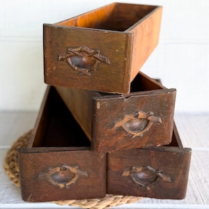 May include: A stack of three rectangular wooden drawers with a dark brown finish. Each drawer front features a carved, centered handle detail with a bow-like design. The wood grain is visible, showing signs of age and wear on the surfaces.