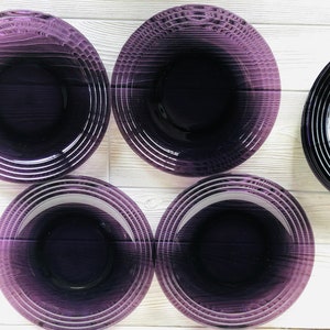May include: Five purple glass plates with a concentric circle design. The plates are arranged in a circle on a white wooden surface.