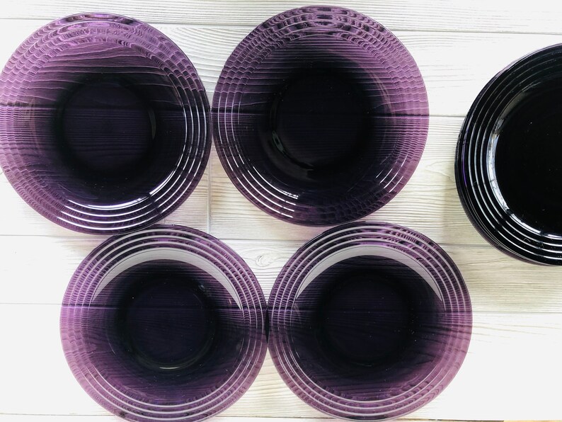 May include: Five purple glass plates with a concentric circle design. The plates are arranged in a circle on a white wooden surface.