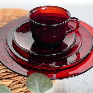 May include: A set of deep red translucent glass dinnerware consisting of a cup, a saucer, and two stacked plates. The items feature a smooth, glossy finish and a uniform ruby color, with the cup positioned centrally on the smaller saucer.