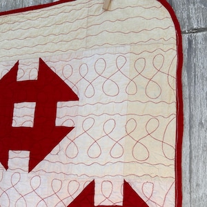 Peut inclure: Une courtepointe faite &agrave; la main avec un motif rouge et blanc. La courtepointe pr&eacute;sente de grandes formes de fl&egrave;ches rouges et des volutes brod&eacute;es rouges sur un fond blanc. Les bords sont bord&eacute;s de tissu rouge. La courtepointe est suspendue.