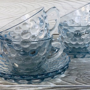 May include: Set of four blue glass teacups and saucers with a textured bubble design. The cups and saucers are stacked on top of each other.