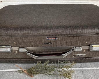 May include: A vintage brown suitcase with a textured exterior, silver hardware, and a handle. The suitcase has a rectangular shape with rounded corners and a metal clasp closure. A sprig of greenery rests in front of the suitcase.