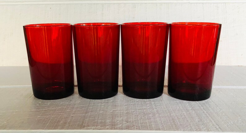May include: Four red drinking glasses. The glasses are cylindrical with a dark red base that fades to a lighter red at the top. The glasses are made of glass and are empty.