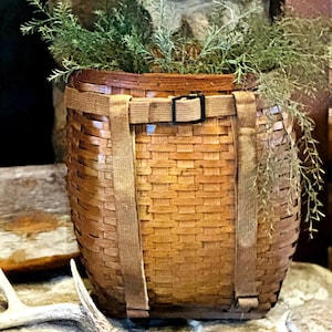 May include: A woven brown basket with a rectangular shape, featuring a buckle closure and tan straps. The basket is filled with green foliage. The basket is sitting on a stone surface.