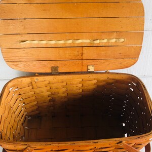 Puede incluir: Una cesta de picnic de madera vintage con un interior de mimbre tejido y una tapa con bisagras. La cesta tiene dos asas de cuero y est&aacute; abierta, revelando el interior vac&iacute;o.