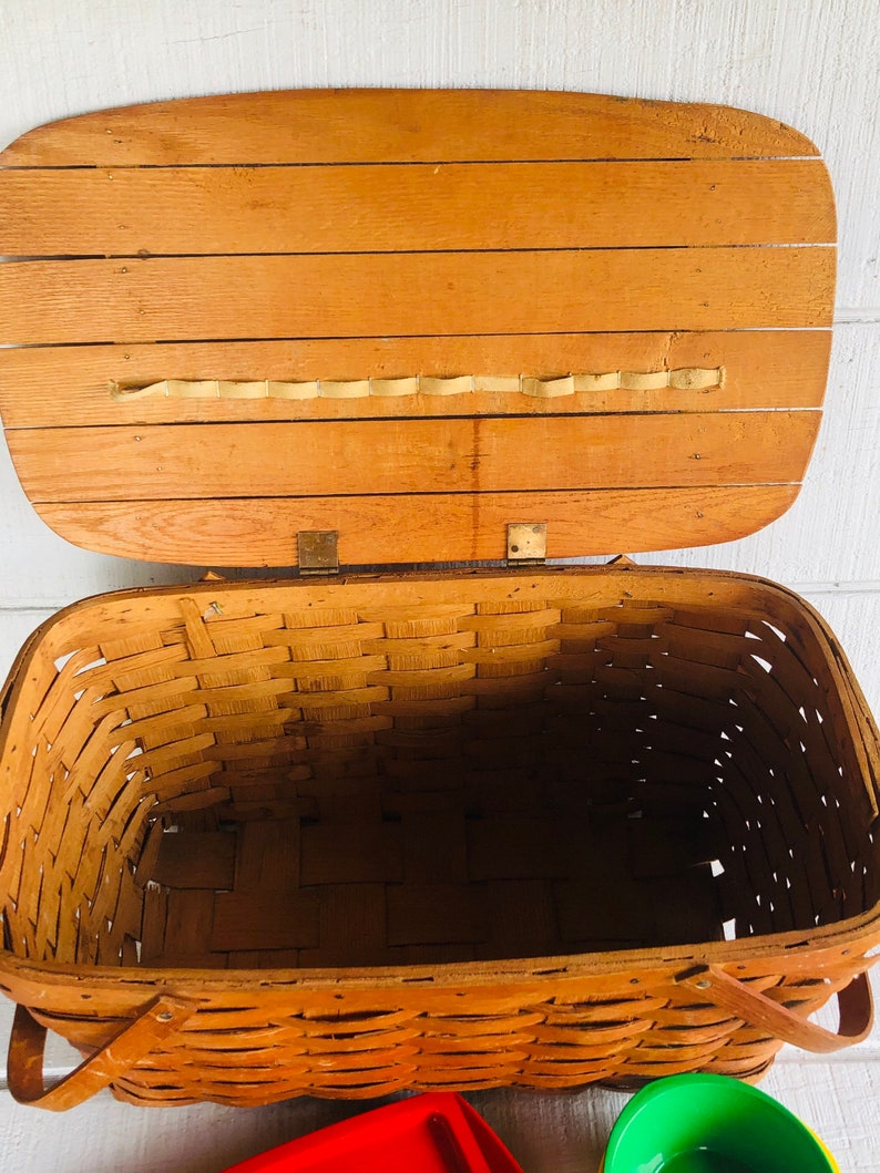 Puede incluir: Una cesta de picnic de madera vintage con un interior de mimbre tejido y una tapa con bisagras. La cesta tiene dos asas de cuero y est&aacute; abierta, revelando el interior vac&iacute;o.