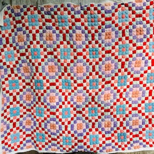 May include: A handmade quilt with a checkerboard pattern in red, white, and various pastel colors. The quilt features raised, textured squares in shades of pink, blue, and purple, creating a dimensional effect. The quilt is hanging against a wooden background.