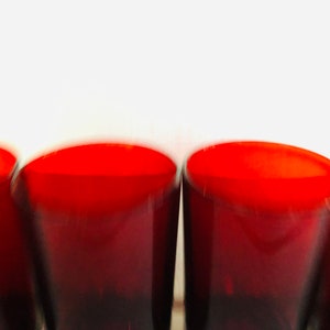 May include: A close-up shot of four red-tinted drinking glasses. The glasses have a rounded, curved shape and are arranged in a row. The top of the glasses are a bright red, fading to a darker red at the base. The background is white.