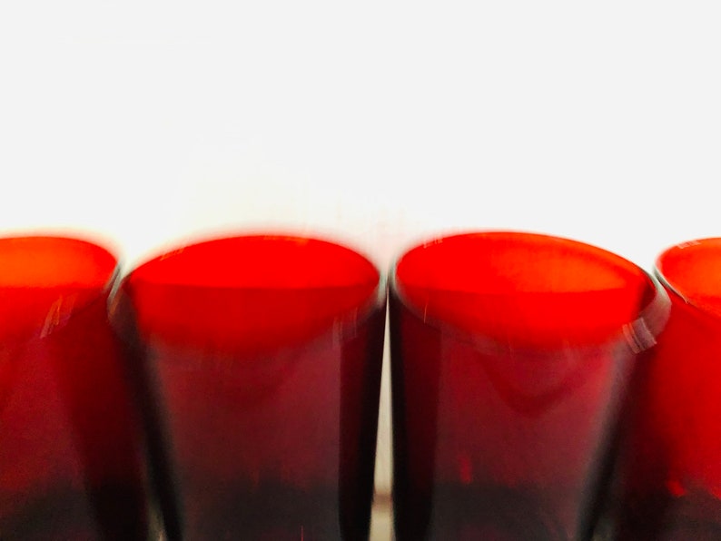 May include: A close-up shot of four red-tinted drinking glasses. The glasses have a rounded, curved shape and are arranged in a row. The top of the glasses are a bright red, fading to a darker red at the base. The background is white.