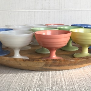 May include: A collection of vintage glass dessert cups in various pastel colors, including blue, white, green, yellow, and pink. The cups are arranged on a wooden tray.