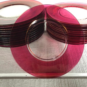 May include: A stack of 20 deep red glass dinner plates with a gold rim. The plates are stacked in a circle, with one plate lying flat in front of the stack.