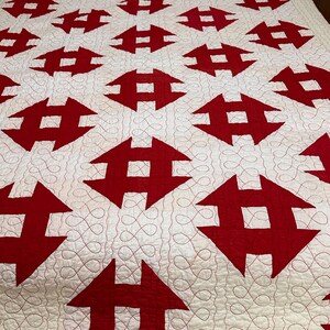 Peut inclure: Une courtepointe faite &agrave; la main avec un motif r&eacute;p&eacute;titif de formes g&eacute;om&eacute;triques rouges sur un fond blanc. La courtepointe est matelass&eacute;e avec du fil rouge, cr&eacute;ant une surface textur&eacute;e. Deux oreillers d&eacute;coratifs blancs sont visibles en haut du lit.