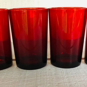 May include: Four tall, red-tinted drinking glasses. The glasses have a gradient effect, transitioning from a deep red at the base to a brighter red at the top. The glasses are cylindrical and appear to be made of glass.