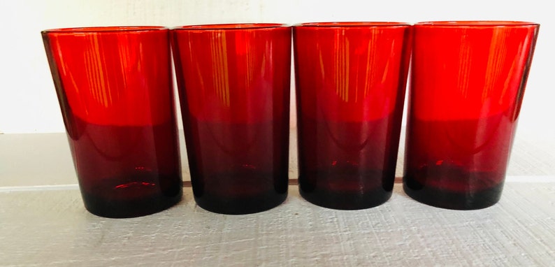 May include: Four tall, red-tinted drinking glasses. The glasses have a gradient effect, transitioning from a deep red at the base to a brighter red at the top. The glasses are cylindrical and appear to be made of glass.