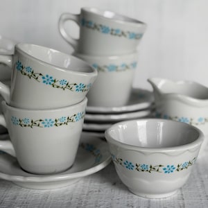 May include: A collection of white ceramic teacups, saucers, and a creamer pitcher, each decorated with a repeating pattern of small blue flowers and thin green vines. The set includes multiple stacked cups and saucers with curved handles.