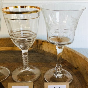 May include: Two clear glass wine glasses with gold trim on a wooden surface. The glass on the left has a gold rim and a textured stem. The glass on the right has an etched floral design. The glasses are labeled "L" and "M".