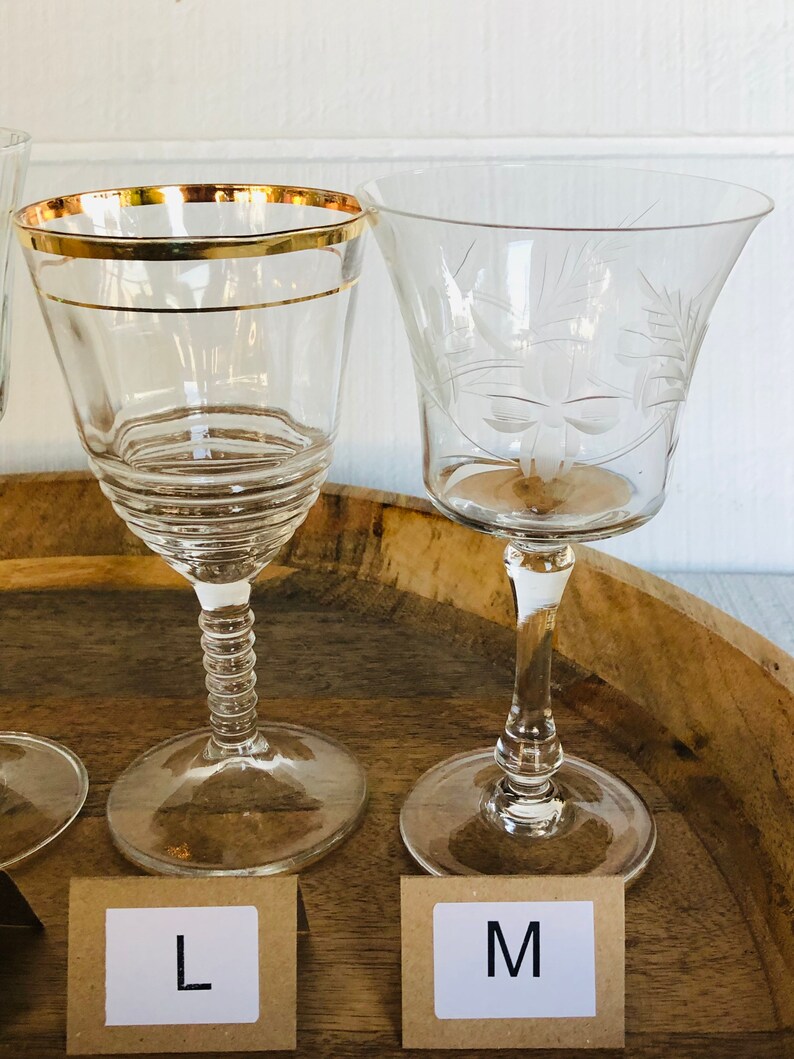 May include: Two clear glass wine glasses with gold trim on a wooden surface. The glass on the left has a gold rim and a textured stem. The glass on the right has an etched floral design. The glasses are labeled "L" and "M".