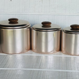 May include: Set of three silver canisters with brown wooden lids. The canisters are arranged in descending size order from left to right.