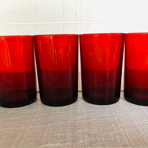 May include: Four red-tinted drinking glasses. The glasses are cylindrical with a slight taper and a dark red base that fades to a brighter red at the top. They are arranged in a row, showcasing their uniform design and color.