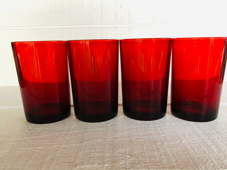 May include: Four red-tinted drinking glasses. The glasses are cylindrical with a slight taper and a dark red base that fades to a brighter red at the top. They are arranged in a row, showcasing their uniform design and color.