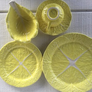 May include: A set of yellow ceramic tableware with a lemon slice design. The set includes two plates, a saucer with a teacup, and a leaf-shaped dish. The plates and saucer have a textured surface, and the teacup has a handle.