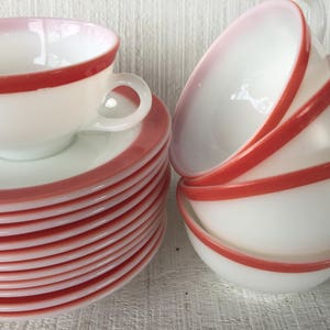 May include: A stack of white saucers and a set of white teacups with a red rim. The teacups are stacked together, and one cup is on top of a stack of saucers. The cups have handles.