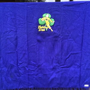 K&ouml;nnte beinhalten: Eine blaue Wolldecke mit einem gestickten Aufn&auml;her, der eine Comicfigur zeigt, die einen Fu&szlig;ball h&auml;lt, und den Text "Fighting Irish". Der Aufn&auml;her ist gelb und gr&uuml;n.