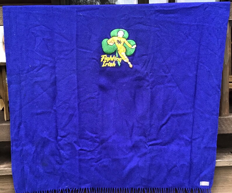 K&ouml;nnte beinhalten: Eine blaue Wolldecke mit einem gestickten Aufn&auml;her, der eine Comicfigur zeigt, die einen Fu&szlig;ball h&auml;lt, und den Text "Fighting Irish". Der Aufn&auml;her ist gelb und gr&uuml;n.