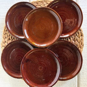May include: A set of five circular plates featuring a deep brown glaze and a prominent concentric ring pattern etched into the surface. Each plate has a smooth, glossy finish and a slightly raised rim, creating a uniform appearance across the collection.