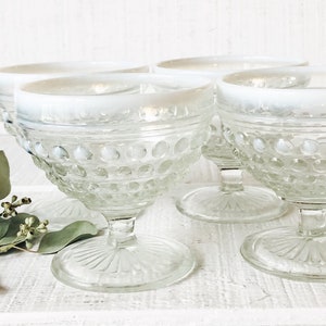 May include: Four clear glass dessert bowls with a white rim and a textured, dot-like pattern. The bowls sit on a clear, stemmed base with a sunburst design. Greenery is placed to the left of the bowls.